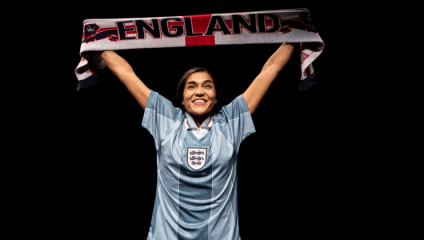 A woman is smiling broadly while holding a scarf above her head that reads ENGLAND. She is wearing a blue England football jersey with the national team's emblem in the center. The black background contrasts with the illuminated subject.