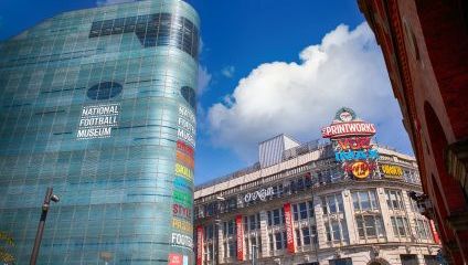 An urban scene showing the National Football Museum, a modern glass-covered building, next to The Printworks, a historic building adorned with colorful neon signs. Both structures contrast against a vibrant blue sky with scattered clouds.