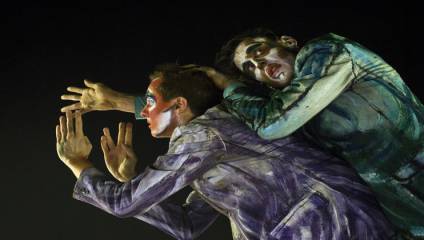 Two performers with faces painted in vibrant colors and eerie designs pose dramatically against a dark background. Both wear costumes with swirling blue and purple patterns. One person holds their hands out in expressive gestures, while the other leans closely behind.