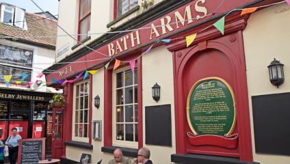 A lively street scene showcases the exterior of Bath Arms pub, adorned with colorful string lights and bunting. Three people are seated at a table outside the pub, nearby is a jeweller's shop with a vibrant display window. The atmosphere is festive and inviting.