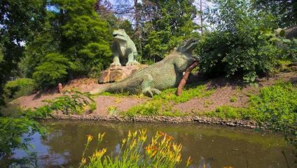 Two large dinosaur sculptures stand among lush greenery by a pond. One dinosaur, posed on all fours, rests on a slope while the other stands more upright, looking over the scene. Bright yellow flowers bloom along the pond's edge, adding color to the landscape.