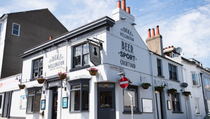 A white-painted pub named The Duke of Wellington stands on a sunny street corner. The building features flower boxes, a Welcome sign, and advertisements for beer and sport in its courtyard. A no-entry sign is visible at the corner. Other buildings are in the background.