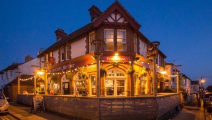 A large, cozy-looking pub or restaurant with warm lighting and festive decorations is seen glowing at dusk. The building has a red and white facade, multiple windows, and a fenced-in outdoor seating area. A sign reads The Ancient Mariner above the entrance.