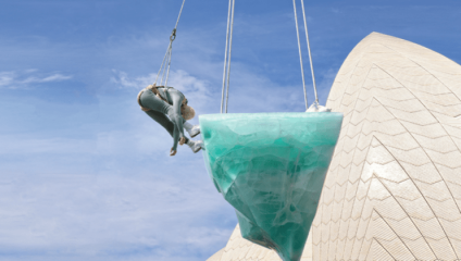 A person in a teal costume is suspended mid-air next to a large, similarly colored, semi-transparent geometric sculpture. They appear to be performing an aerial act against the backdrop of a modern, architecturally distinctive building with a cream-colored facade.