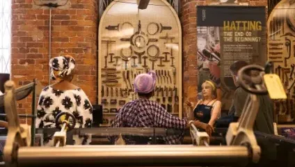 Three people are engaged in conversation in front of a wall display featuring an array of vintage tools. One woman, in work attire, explains something to two others, who are wearing colorful, patterned headscarves. The setting is inside an industrial-themed museum.