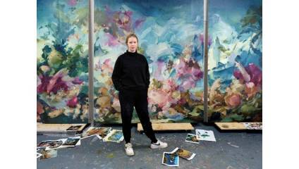 A person stands in front of a large, colorful abstract painting wearing a black hoodie and pants. The floor around them is scattered with various smaller artworks and papers. The background features vibrant hues of pink, blue, and green.