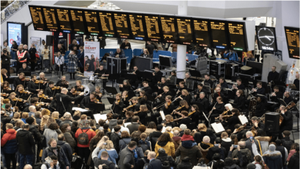 A large crowd gathers around an orchestra performing at a busy train station. The electronic departure board displays numerous train schedules above the musicians. The scene is bustling with activity, as people capture the event with their phones and cameras.