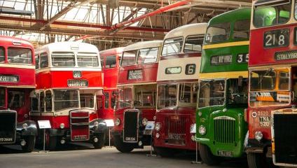 A row of vintage double-decker buses in various colors, including red, green, and cream, are lined up inside a spacious warehouse with a high ceiling and metal beams. The buses display different route numbers and destination signs on their fronts.
