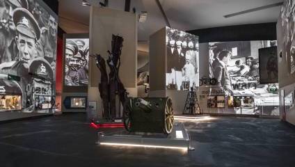 A museum exhibit showcasing artifacts and large historical photographs. Prominently displayed are a military uniform, old artillery, and vintage equipment. The walls feature enlarged black and white photos of military personnel and significant historical moments.