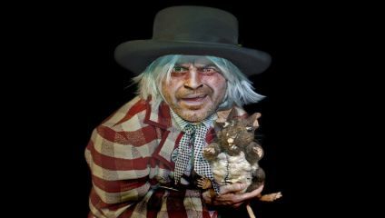 A man in a wide-brimmed hat and checkered attire holds a small, dark-colored animal, possibly a rat. He has disheveled, white hair and a weathered face, and appears to be in a theatrical or dramatic setting against a black background.