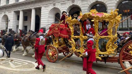 A lavish, gold-adorned carriage is being pulled by horses through a city street during a parade. People in red and gold ornate costumes escort the carriage. The surrounding architecture includes grand, arched windows and columns. Onlookers are visible in the background.
