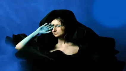 A person with a blue-painted hand and blue eye makeup poses against a blue background. They are wrapped in voluminous, black fabric, creating dramatic folds around their shoulders. The person gazes directly at the camera with their fingers slightly splayed over their eyes.