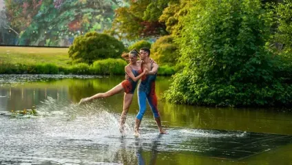 Three dancers perform on a water-covered surface surrounded by lush greenery. One dancer in red leans backward with a leg lifted high, supported by two dancers in colorful outfits, creating a dynamic and captivating scene with splashing water effect.
