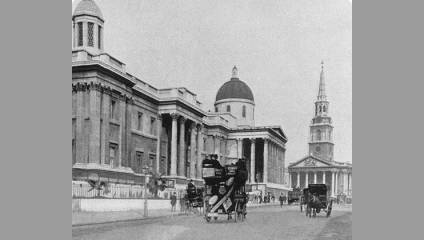 Black and white photo of a historical city street featuring vintage horse-drawn carriages and early double-decker buses. Prominent structures, including a domed building and a tall, narrow church steeple, line the side of the street. Pedestrians are visible in the background.