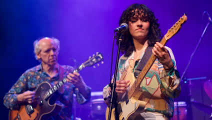 A woman with curly hair and a colorful shirt plays an electric guitar and sings into a microphone on a stage. Next to her, an older man in a patterned shirt plays another electric guitar. They perform under purple stage lights with a drum set visible in the background.
