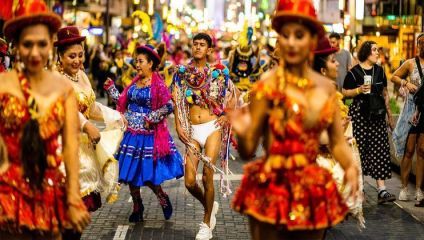 A vibrant street parade featuring dancers in colorful traditional costumes. The participants wear elaborate outfits adorned with sequins and fringe, with a diverse crowd watching from the sides. Bright city lights illuminate the lively scene.