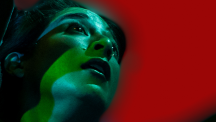 Close-up of a person's face illuminated with green light, against a red background. The person appears to be looking upwards with a curious or contemplative expression. The lighting casts shadows that accentuate the contours of the face.