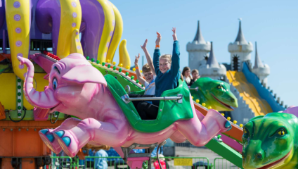 Children enjoy a ride on a colorful, elephant-themed amusement ride at a fair. The girls have their arms raised in excitement. In the background, there are other vibrant rides and slide structures resembling a castle under the clear blue sky.