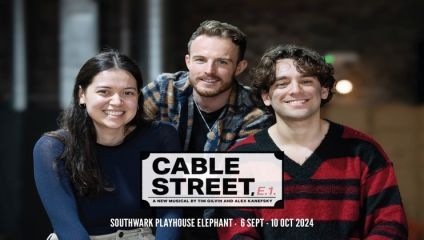 Three people are smiling and posing together in a relaxed setting. The text in the image reads: Cable Street, E.1. A new musical by Tim Gilvin and Alex Kanefsky. Southwark Playhouse Elephant. 6 Sept - 10 Oct 2024. One person is wearing a striped sweater.