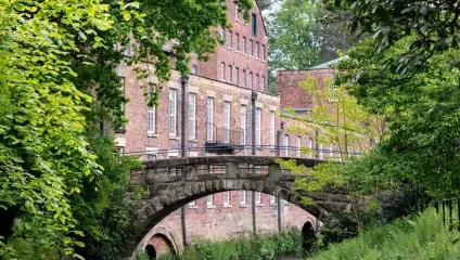 A serene brick building, partially obscured by lush green foliage, stands alongside a stone arch bridge over a narrow waterway. The scene is quiet and picturesque, emphasizing the natural and historic elements harmoniously coexisting.
