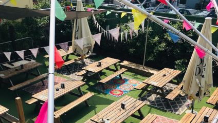 Outdoor seating area with wooden picnic tables and benches set on artificial grass, decorated with colorful string lights and bunting. The area is shaded by umbrellas and surrounded by greenery, creating a festive, inviting atmosphere.