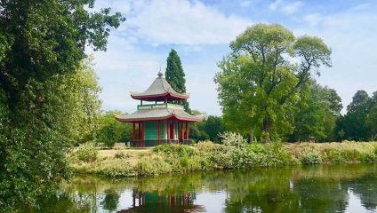 A traditional Asian-style pavilion with a multi-tiered roof is situated among lush greenery by the edge of a calm lake. Tall trees surround the structure, and the sky above is partly cloudy with blue patches. The pavilion is reflected in the water.