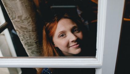 A person with long red hair is looking up at the camera while standing by a window. They are partially obscured by the window frame and curtain, and they have a subtle smile on their face.