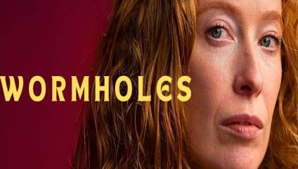 A close-up of a person with red hair and light eyes looks pensively at the camera against a maroon background. The word WORMHOLES is written in large yellow letters to the left of the person's face.