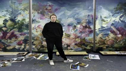 A person in a black outfit stands confidently in an art studio. The studio is filled with vibrant, abstract paintings of natural scenery. Artistic works and papers are scattered on the floor around them. Large canvases with colorful, nature-inspired art adorn the walls.