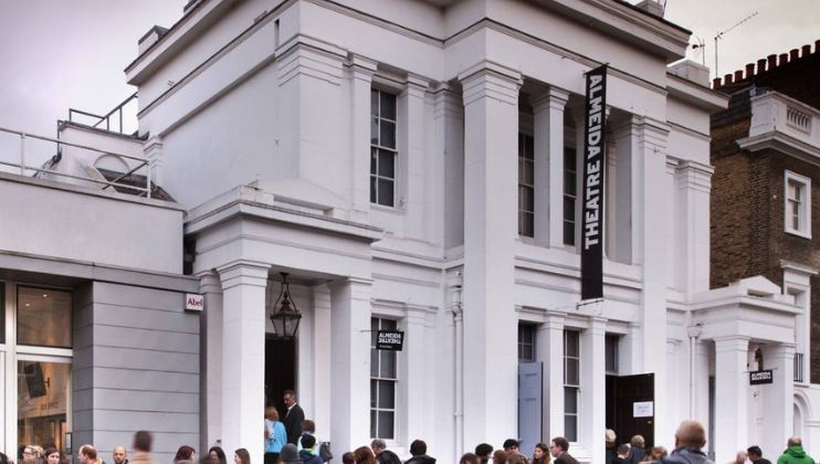 A crowd of people gathers outside a white, columned building with a sign reading THEATRE on a vertical black banner. The building has a classical facade and large windows. The scene appears lively, suggesting an event or performance is taking place.