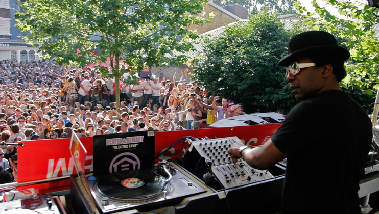 A DJ is performing on an outdoor stage, surrounded by electronic equipment, with a large, enthusiastic crowd gathered below. The DJ is wearing sunglasses and a black hat. Trees and buildings are visible in the background, contributing to the lively atmosphere.