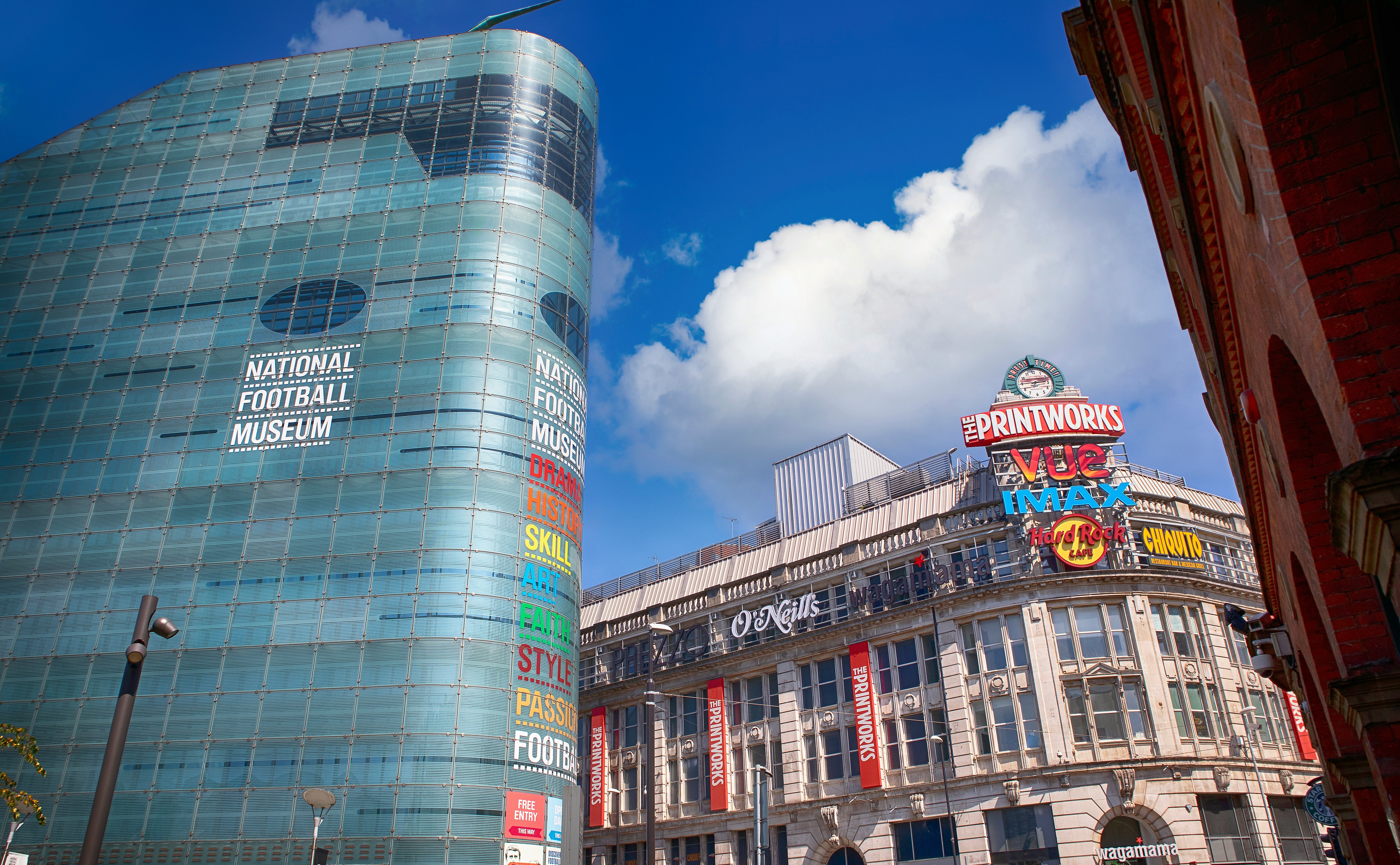 An urban scene showing the National Football Museum, a modern glass-covered building, next to The Printworks, a historic building adorned with colorful neon signs. Both structures contrast against a vibrant blue sky with scattered clouds.