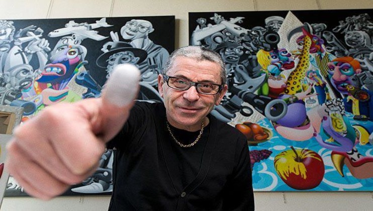 A person wearing glasses and a black sweater gives a thumbs-up to the camera. Two vibrant, surrealist artworks are displayed on the wall behind them, featuring colorful characters and imaginative scenes. The individual appears to be in an art gallery or studio.