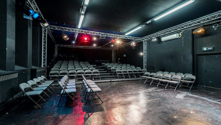 A small theater with tiered seating. The room has black walls and ceiling with exposed metal framework and stage lighting. Rows of folding chairs are arranged facing an open stage, with additional raised seating in the back.