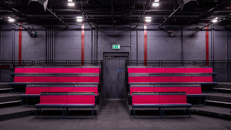 A modern, empty theater with tiered seating featuring vibrant pink chairs. The walls are dark gray, and electrical wiring is exposed on the ceiling. There is an exit door at the back center of the seating area with a green Exit sign above it.