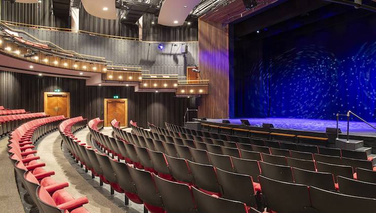 A theater auditorium with rows of red and black seats curving around towards a stage. The stage features a blue backdrop with star-like patterns. The walls have vertical lines, and there are several doors and lights visible. The theater is empty.