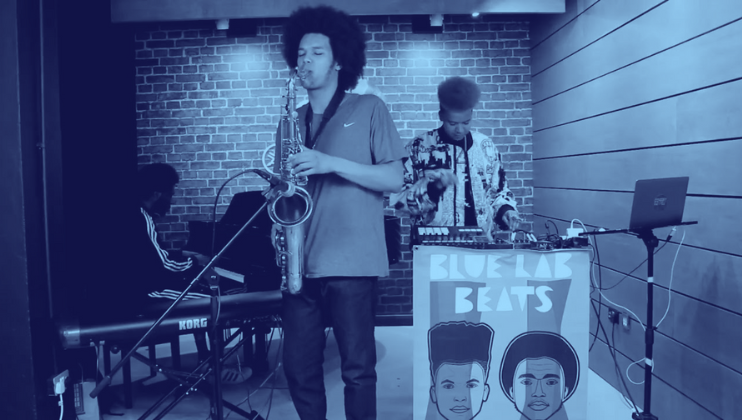 A musical trio performs in a blue-lit studio. The first musician plays the saxophone, the second operates a keyboard, and the third handles a mixing console at a station with a Blue Lab Beats sign featuring illustrated faces. A brick wall backdrop adds to the ambiance.