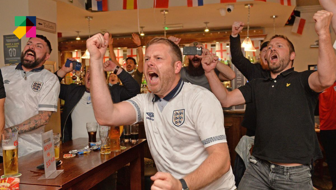 A group of ecstatic soccer fans in a pub, adorned with flags, celebrate a goal. The fans, wearing team jerseys, cheer with fists raised in the air. The atmosphere is joyous and vibrant, with drinks on the table and one fan capturing the moment on a phone.