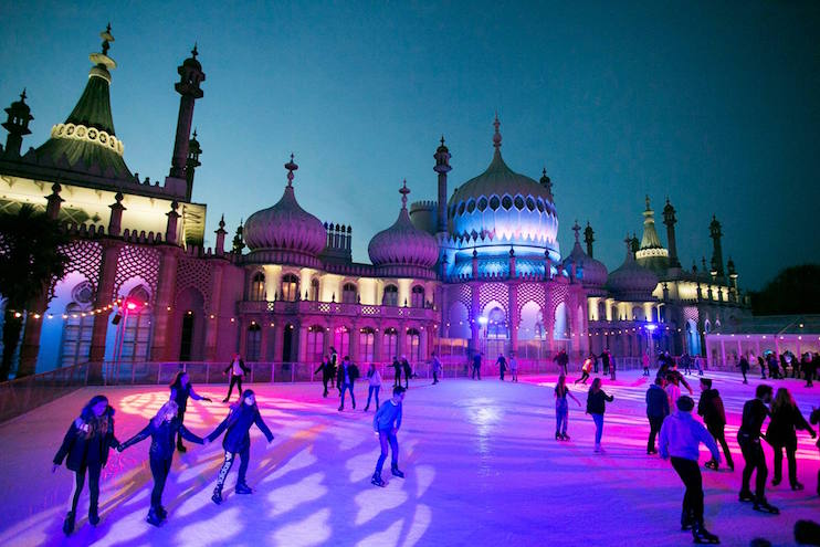 People are skating on an ice rink illuminated by colorful lights at dusk. The background features a grand building with ornate domes and towers, reminiscent of Indian architecture. The sky above is a deepening blue, enhancing the vibrant ambiance.