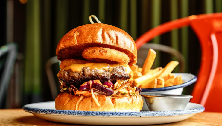 A juicy cheeseburger with a thick patty, cheese, slaw, and an onion ring in a toasted bun is presented on a plate next to a side of crispy French fries and a small metal cup of dipping sauce. The background shows blurred cafeteria-style seating.