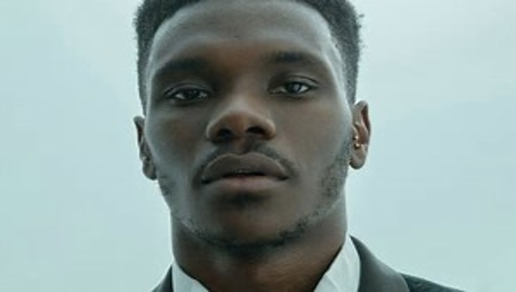 Close-up of a young person with short, curly black hair and an earring in their left ear. They are dressed in a white shirt and a dark blazer, with a neutral expression, standing against a light, blurred background.