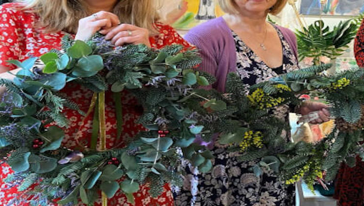 Two women are holding wreaths decorated with green leaves, eucalyptus, and small red berries. The woman on the left is dressed in a red floral dress, and the woman on the right is wearing a purple sweater over a black and white floral dress.