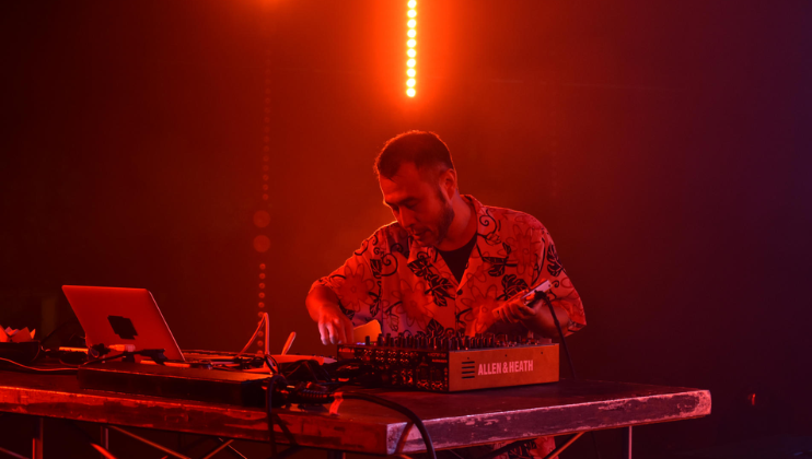 A man in a patterned shirt operates a sound mixer on a dimly lit stage with red lighting. A laptop and various cables are also on the table. Vertical light beams in the background add to the ambiance.