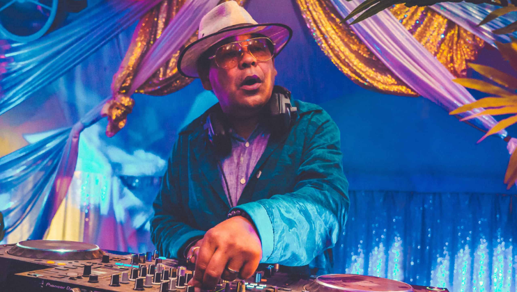 A DJ wearing a light-colored hat, round sunglasses, headphones, and a teal jacket is playing music on a mixing console. The background is decorated with colorful fabric drapes in blue and purple tones, along with some golden accents and palm leaves.