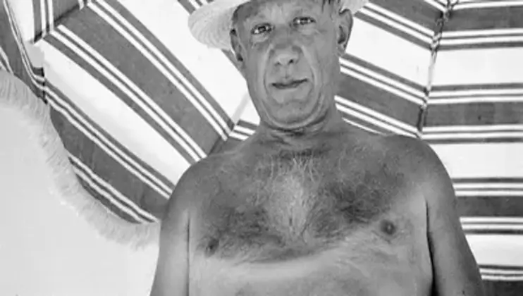 A shirtless man wearing a straw hat stands in front of a striped fabric background, possibly an umbrella or tent. The image is black and white.