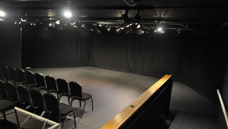 A small black box theater with dim lighting. The stage is empty, with black curtains in the background. Several rows of black chairs are positioned in the audience area, facing the stage. The setting is intimate, ideal for small performances.