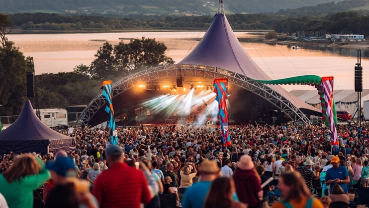 A large crowd enjoys an outdoor music festival at dusk, gathered in front of a brightly lit stage under a tent-like structure. Scenic mountains and a lake are visible in the background. Colorful banners and smaller tents are dispersed throughout the festival area.