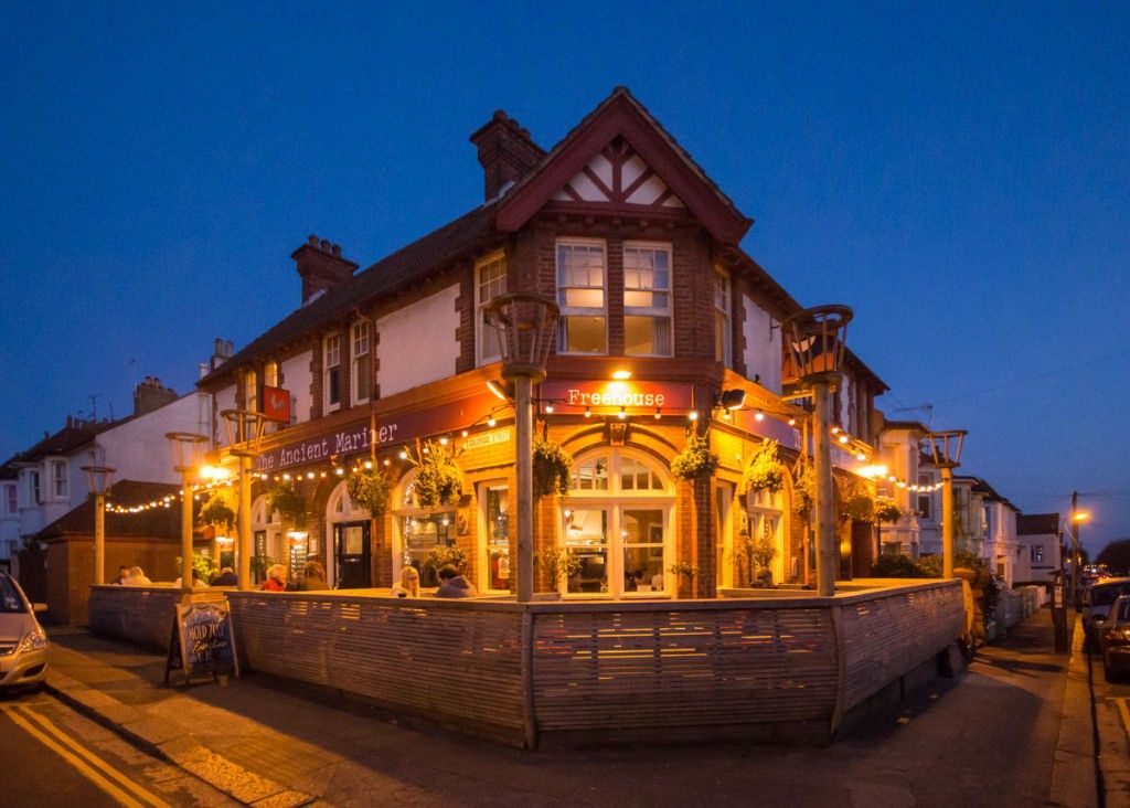 A large, cozy-looking pub or restaurant with warm lighting and festive decorations is seen glowing at dusk. The building has a red and white facade, multiple windows, and a fenced-in outdoor seating area. A sign reads The Ancient Mariner above the entrance.