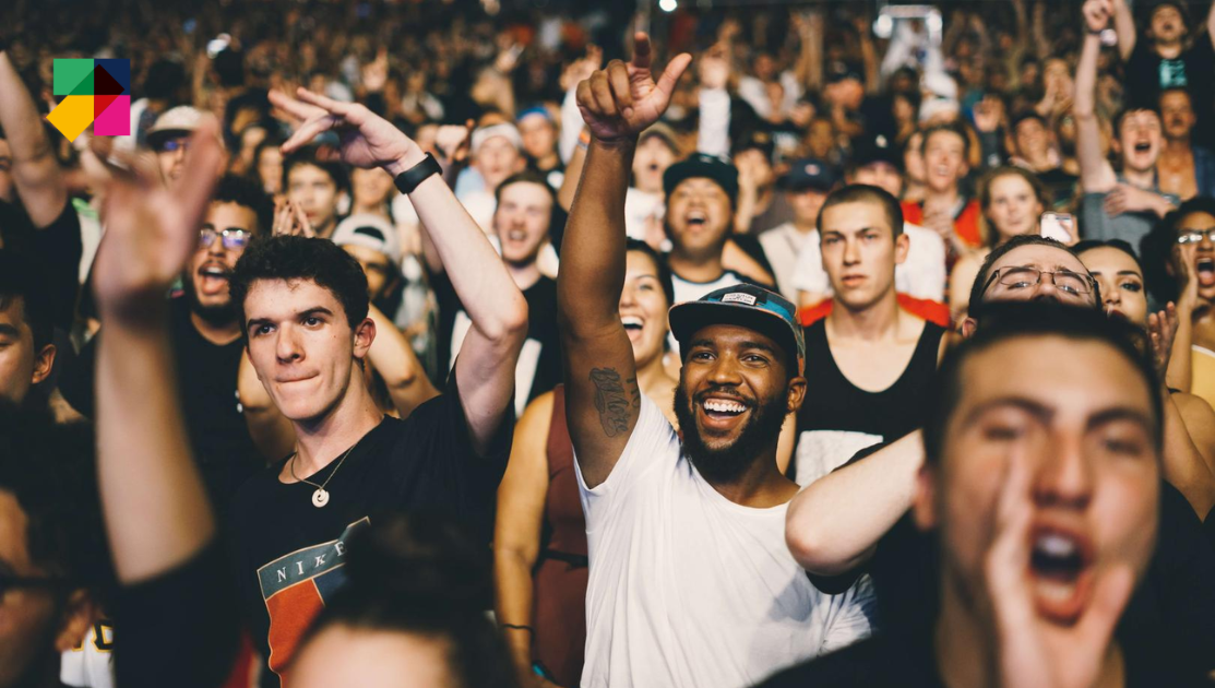 A large, diverse crowd of people are at a concert or event, excitedly cheering and raising their hands. The atmosphere is energetic and joyful. In the foreground, individuals appear animated and cheerful. A colorful logo is visible in the upper left corner.