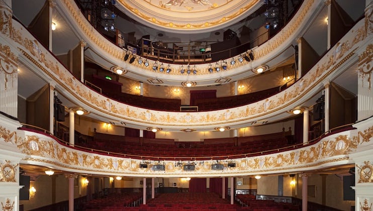 A view of an ornate theater's interior from the stage, showing three tiers of seating adorned with intricate gold detailing, red upholstered seats, and a chandelier hanging from the ceiling in the center. The theater is illuminated by stage lights aimed at the audience area.